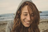 An auburn haired, freckled woman wearing a grey knitted hoodie. She’s on a beach, smiling at something down and away from the camera lens.