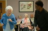 A senior in a senior-center environment dances next to a young adult