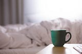 steaming cup on a desk in front of a soft bed and blanket
