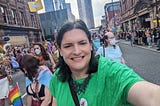 Chris Northwood, smiling, with a trans flag on her cheek, walking down Deansgate with towers in the background and crowds onlooking with a group of others around her