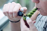 Person holding a green weed pipe in the mouth while lighting cannabis leaves in it.