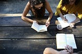 group of three people reading together and taking notes outdoors at picnic table