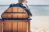 Sitting on a wooden deck chair looking out to sea with chin in hand, waiting.