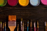 An image showing five paint colors at the top, and different size paint brushes at the bottom on a dark wooden table.