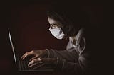 A woman wearing a mask typing on a laptop in the dark