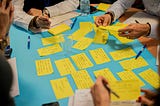 A group of people around a pale blue table are writing and exchanging yellow post it notes, a writers workshop.