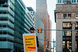 Photo of a man stood on a street in a city, holding a yellow sign which reads “slavery still exists”.