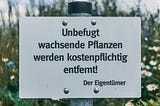 White sign on metal pole in flowery summer garden, in German, reading “Unbefugt wachsende Pflanzen werden kostenpflichtig entfernt! Der Eigentümer”