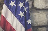 A close up of a partially-folded American flag, with the creases in its canvas visible, in front of blurred gray stone wall.