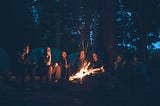 Several people sitting around a campfire in the woods at night.