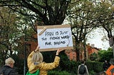 A photo of a woman standing along a race course, holding aloft a sign that says, “Pain Is Just the French Word for Bread”