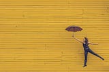 A woman jumping in front of a yellow wall while holding an umbrella.