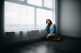 A woman is sitting on the floor near the window of an empty apartment. Anthony Tran/Unsplash.