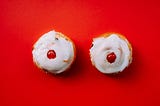 Two vanilla cupcakes with red cherries in the center, meant to look like breasts, set against a red backdrop.