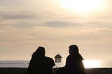 Two women talking to each other by a lake or a sea and a wooden house built on the sea or lake is visible between them.