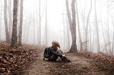 Winter forest, muted colors, girl with daypack sits on trail facing to the side; her hair blowing across covers her face