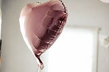 A pink, heart shaped balloon floating in a white walled room.