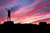 A photograph of a man on a cliff with his hands up in the air reaching towards the sky facing a sunset