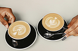 A close-up image of two hands holding black mugs filled with latte