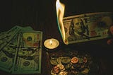 A hand holding a burning $100 bill above a pile of coins and other bills next to a lit candle.