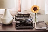 A typewriter, a white lamp, a sunflower in a vase, and a notebook