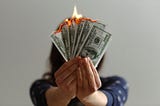 A person wearing a blue shirt holds several hundred dollar bills in a fan shape, covering her face. The bills have been lit on fire.