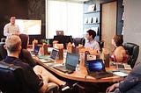 A group of employees are in a meeting around a table. Each one has their own laptop except for one person who’s standing by a larger screen speaking to them all.