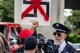 A man in a uniform holds up a sign with a swastika, crossed out in red paint, as a crew films him