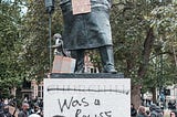 An image of a group of people in protest against a Winston Churchill statue that has been spray painted with words to read “Churchill was a racist”. The statue holds up a sign that reads “Black Lives Matter”