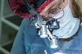 A close-up of a surgeon wearing magnifying loupes, a headlamp, surgical mask, and red cap while performing a procedure. The focused lighting highlights the precision tools and the surgeon’s intense concentration.