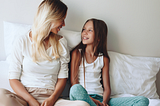 Woman sitting on a bed smiling down at her tween daughter.