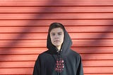 a youth in a dark hoodie standing against a reddish wall