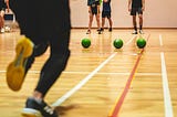 Indoor court, person in track-suit running towards three balls. In the background. three players waiting.