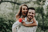 A picture of a happy couple, a man and a woman. The woman has jumped up onto her husband’s back and is holding a rose.