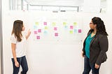 Two women collaborating at a whiteboard with sticky notes organised into workflow columns, discussing iterative delivery in a bright modern office