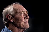 An old man with white hair, wrinkly face, and chequered shirt looking to the right