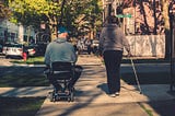 A photo of a man in an electric mobility scooter and a woman walking with a cane. The man is wearing a grey hoodie, black sweatpants with white stripes, and a blue baseball cap. The woman is wearing a grey hoodie, with matching grey sneakers, and dark pants. She has medium brown hair. Her cane is white with a pattern of black circles. The photo is taken of the couple’s back as they travel next to one another down a city sidewalk.