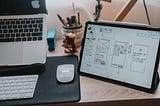 A Macbook, a keyboard, a mouse and an iPad displaying handrawn wireframes are on a wooden desk.