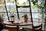 A small round table with 2 chairs by a window. On the table is a French press, coffee cups, and a potted plant. There are trees arching overhead.