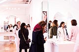 Customers browse a high-end looking cosmetics store with bright white lights as sales associates approach them to help with their shopping needs.