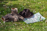 A pair of boots, a hat, a map, and a compass lying on grass dappled with small white flowers