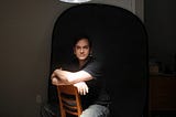 A man with dark hair & pale skin sits backwards on a chair on a dark room, lit dramatically from above.