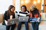 Women sitting on a couch, working together on their laptops.