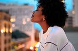 Head-to-shoulders, side shot of pretty lady with afro, eyes closed, head tilted upward, standing on a balcony that overlooks city lights.