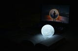A glowing moon lamp laying on an open book, laptop in background
