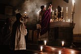 A priest elevates the Host for Communion in a very smoky, candle-lit chapel as two other clergy look on. One of them might be a deacon.