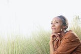 A Black woman, sitting in a grassy field and looking pensive.