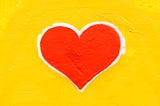 Red heart, with a white outline, painted on a bright yellow, stucco, wall