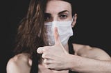 A woman in a blue disposable face mask makes a thumbs-up gesture.