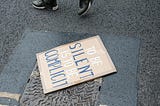 a sign stating “to be silent is to be complicit” lays on the ground